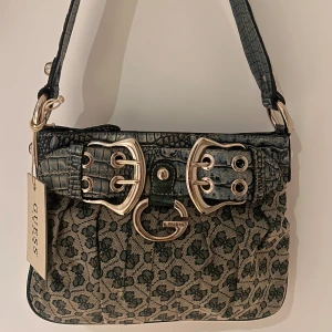 Guess axelväska med mönster och spännen - Cool axelväska från Guess med beige och mörkgrönt monogrammönster, detaljer i krokopräglat syntetmaterial och stora silvriga spännen framtill. Väskan har dragkedja bak och insida med leopardmönstrat foder. Perfekt för dig som gillar statement-accessoarer.