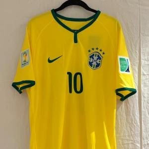 Brasilien Neymar JR VM 2014 tröja - En retro Brasilien fotbollströja från VM 2014 med Neymar JR och nummer 10 på ryggen. Tröjan är gul med gröna detaljer, har korta ärmar och är tillverkad i Nike Dri-Fit material. Brasilien märker är broderat på bröstet.