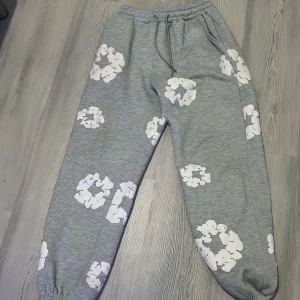 Grå mjukisbyxor med vita molnmönster - Grå mjukisbyxor från Denim Jeans med vita molnliknande mönster över hela byxorna. De har elastisk midja med snörning och muddar vid bensluten för extra komfort. Perfekt för chill dagar och avslappnad stil.