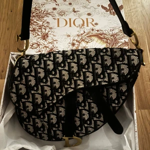 Svart saddle bag - Saddle axelväska med svart och beige monogrammönster. Väskan har en unik sadelform, guldfärgade metalldetaljer med CD-logga och ett brett axelband i svart. Köpt här på Plick för 1500 kr för något år sedan. Använd bara några gånger och är i toppenskick.