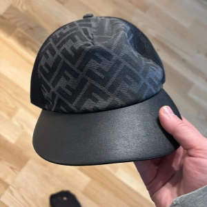 Svart Fendi keps med mönster - Snygg svart keps från Fendi med ikoniskt FF-mönster på ovansidan och stilren, slät skärm i skinn. Perfekt accessoar för dig som gillar exklusiv streetstyle och vill sticka ut med designerplagg.