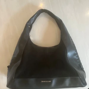 Svart axelväska från Riversvart Island - Snygg svart axelväska från River Island med mjuk form och bred axelrem. Väskan har detaljer i skinn och mocka, guldfärgad dragkedja och flätad kant på sidan. Perfekt storlek för allt du behöver och stilren look.