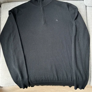 Svart half zip stickad tröja JLindeberg - Svart stickad tröja från J.Lindeberg med half zip och diskret logga på bröstet. Tröjan har hög krage, ribbade muddar och är långärmad. Perfekt för lager-på-lager och snygg till både jeans och chinos.