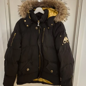 Svart Moose Knuckles dunjacka med päls - Svart dunjacka från Moose Knuckles med guldfärgade detaljer och dragkedjor. Skavanker finns som syns på bilderna. Där av priset.
