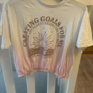 Vit och rosa croppad t-shirt med tryck - Säljer en croppad t-shirt i vitt och ljusrosa med soltryck och texten 'Creating goals for me'. Toppen har korta ärmar, rund hals och dragsko nertill för en snygg passform. Perfekt för dig som gillar chill och trendiga vibes.