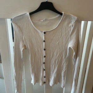 Vit ribbadstickad topp med knappar - Snygg vit ribbad topp med långa ärmar och knappar hela vägen framtill. Toppen har en rund halsringning och är figurnära i modellen. Perfekt att styla med jeans eller kjol för en clean look. (Obs, tröjan går inte att knäppa upp)