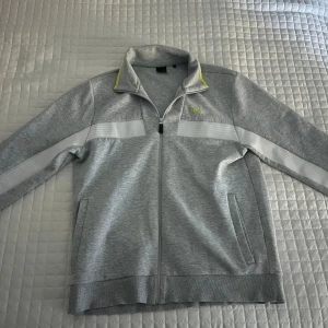 Grå ziptröja från BOSS med vita detaljer - Snygg grå ziptröja från BOSS med vita ränder längs ärmarna och diskret gul BOSS-logga på bröstet. Den är knappast använd nästan helt ny, köptes för 2200kr 