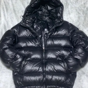 Svart pufferjacka från Zara med huva - Säljer en svart, glansig pufferjacka från Zara med huva och dragkedja. Jackan är vadderad och har en oversized look med långa ärmar och två sidofickor. Perfekt för kalla dagar när du vill hålla stilen och värmen.