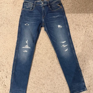 Replay Anbass blå jeans med slitningar - Snygga blå jeans från Replay, modell Anbass, med coola slitningar och tvättade detaljer framtill. Klassisk femficksdesign, gul kontrastsöm på bakfickorna och Replay-lapp i midjan. Jeansen har en smal passform och är tillverkade i stretchigt denim för skön komfort.