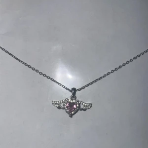 Rosa halsband - Pris kan diskuteras!!Sött halsband i silverfärg med ett hjärtformat hänge. Hjärtat har en rosa sten i mitten och små glittrande stenar runtom, formade som små vingar. Kedjan är tunn och elegant – passar både till vardag och fest. I fint skick! S925 silver