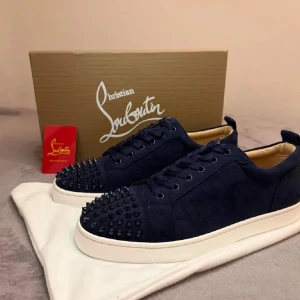 Mörkblå sneakers från Christian Louboutin - Snygga mörkblå sneakers från Christian Louboutin i mocka med vita sulor och ikoniska röda undersidor. Skorna har snörning och coola svarta nitar på tån för extra edge. Insidan är beige och de har en rund tå. Perfekt för dig som vill sticka ut med lyxig streetstyle.