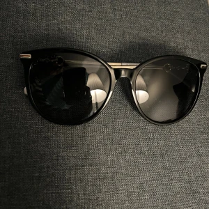Svarta solglasögon från Gucci - Snygga svarta solglasögon från Gucci med rundad form och coola gulddetaljer på skalmarna. Bågarna är i blank plast och metall, med Gucci-logga på sidan. Perfekt accessoar för att lyfta din stil. Pris går att diskutera