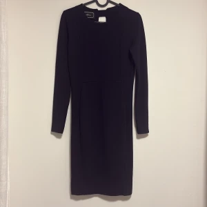 Svart långärmad klänning By Malene Birger - Snygg svart klänning från By Malene Birger med lång ärm och stilren, rak passform. Klänningen har en diskret v-ringning i ryggen och är knälång, perfekt för dig som gillar minimalistisk design. Materialet är mjukt och följsamt polyester.