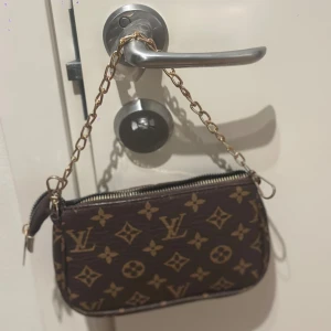 Brun handväska med guldkedja - Klassiskt monogrammönster i beige och guld. Väskan har en guldfärgad kedja som axelrem och dragkedja upptill. Märkeslogga i läder framtill och insidan är svart. 12x19cm