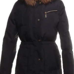 Svart dunjacka med päls från Hollies  - Snygg svart dunjacka från Woolrich med lång passform och fluffig brun päls runt huvan. Jackan har dragkedja och knappar framtill, en bröstficka med dragkedja och markerad midja. Perfekt för kalla vinterdagar när du vill hålla stilen.