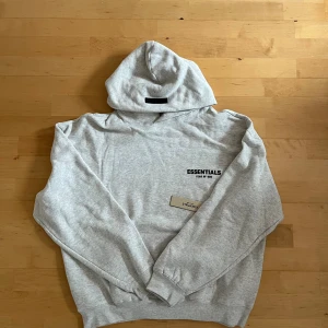 Grå Essentials Fear of God hoodie - Snygg ljusgrå hoodie från Essentials Fear of God med svart logotyptryck både fram och bak. Tröjan har huva, ribbade muddar och en klassisk känguruficka. Perfekt för dig som gillar streetwear och vill ha en clean look.
