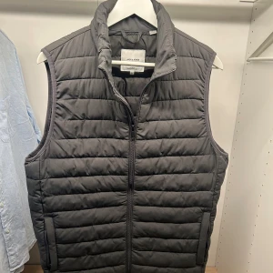 Svart quiltad dunväst Jack & Jones S - Svart dunväst från Jack & Jones i storlek S. Västen har ett quiltat, horisontellt mönster och dragkedja framtill. Hög krage och två sidofickor. Perfekt att slänga över en hoodie för extra värme. Materialet är polyester och dun.