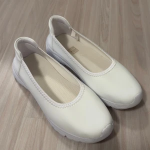 Vita ballerinaskor med platt sula - Stilrena vita ballerinaskor med rund tå och platt, mönstrad sula för extra grepp. Ovandelen är i syntetmaterial och insidan har en mjuk textil. Perfekta för dig som gillar enkel och clean stil.