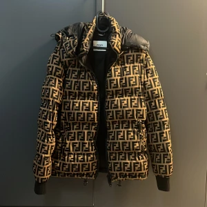 Fendi pufferjacka med Fendi mönster - Säljer en fet pufferjacka från Fendi med ikoniskt FF-mönster i svart och beige. Jackan har huva, ribbade muddar och dragkedja framtill. Dock är dragkleppen av på ena dragkedjan och den andra har lite rost.  Säljs för 5000 kr ( snabb affär kan priset diskuteras) 