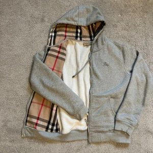  Burberry hoodie - Säljer en grå hoodie från Burberry med klassisk rutig insida i beige, svart och röd. Tröjan har huva med snörning, dragkedja och broderad Burberry-logga på bröstet. Mjuk insida och två fickor framtill. Perfekt för dig som gillar lyxiga streetwear-vibes.