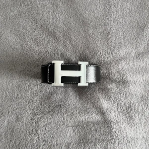Hermès Bälten - Säljer ett stilrent svart Hermès-bälte i skinn med ett ikoniskt H-spänne i silverfärgad metall. Bältet har vit kontrastsöm längs kanterna och en klassisk, rak form. Perfekt för dig som vill ha en lyxig accessoar med tydlig designer-vibe.