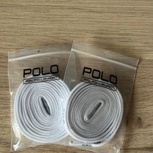 Vita skosnören Polo Ralph Lauren - Säljer ett par vita skosnören från Polo Ralph Lauren, perfekta för att ge dina sneakers en fräsch och clean look. Skosnörena har Polo-loggan tryckt i grönt längs hela snöret och är tillverkade i slitstark syntet. Passar till de flesta sneakers och skor med snörning.