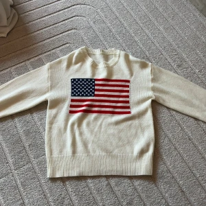 Vit stickad tröja med USA-flagga - Snygg vit stickad tröja med amerikansk flagga framtill. Tröjan har rund halsringning, långa ärmar och ribbade muddar vid ärmslut och nederkant. Perfekt för dig som vill sticka ut med en klassisk och cool look.