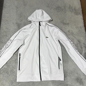 Vit vindjacka med huva från Lacoste - Snygg vit vindjacka från Lacoste med svart dragkedja och huva. Jackan har Lacoste-tryck längs ärmarna och klassisk krokodillogga på bröstet. Tillverkad i polyester med svarta detaljer och fickor med dragkedja. Perfekt för en sportig och clean look.