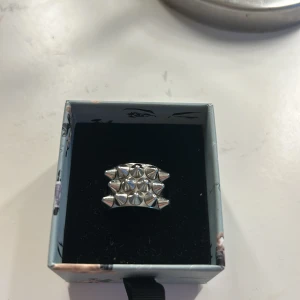 Silvrig ring med nitar och spikes - Cool och edgy silvrig ring med flera små nitar och spikes i ett rutmönster. Ringen har en bred form och ger en riktigt tuff vibe, perfekt för dig som gillar att sticka ut. Passar både till streetwear och rockiga outfits.