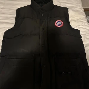 Svart dunväst från Canada Goose - Svart dunväst från Canada Goose med klassisk logga på bröstet. Västen har hög krage, tryckknappar framtill och stora fickor. Insidan har meshficka och slät polyesterfoder. Perfekt för kalla dagar och har en stilren, enkel look.