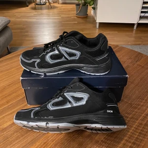 Svarta Dior B30 sneakers med grå detaljer - Säljer ett par svarta Dior sneakers med grå och silverfärgade detaljer. Skorna har en chunky sula i grått och svart, meshpaneler för extra andningsförmåga och tydlig Dior-logga på sidan och sulan. Klassisk snörning och sportig vibe, perfekta för dig som gillar exklusiva streetwear-vibbar.