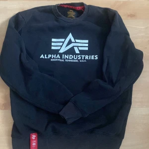 Svart sweatshirt Alpha Industries S - Svart sweatshirt från Alpha Industries i storlek S med stort vitt tryck på bröstet och klassisk rund hals. Tröjan har långa ärmar, ribbade muddar och en röd detalj vid nederkanten. Perfekt för dig som gillar streetwear och snygga loggor.