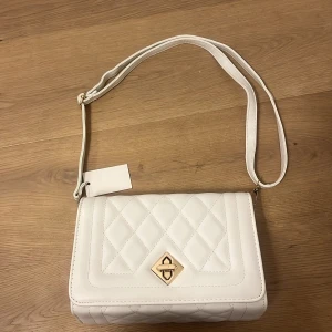 Vit quiltad axelväska med kedja - Snygg vit axelväska med quiltat mönster och guldfärgad vridlåsdetalj framtill. Väskan har både justerbar axelrem och en chunky kedja i vitt och guld som kan användas som handtag. Insidan har fack med dragkedja för extra förvaring. Perfekt för att lyfta din outfit. Helt ny och inte använd. 
