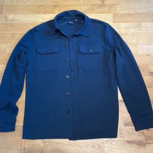 Mörkblå overshirt från Only & Sons  - Snygg mörkblå overshirt från Only & Sons i storlek XS. Skjortan har två bröstfickor med knappar, klassisk krage och knäppning hela vägen. Materialet är mjukt och känns som bomull, perfekt för lager-på-lager. Enkel och stilren design som funkar till många outfits. Den är storlek XS men sitter som en S!!