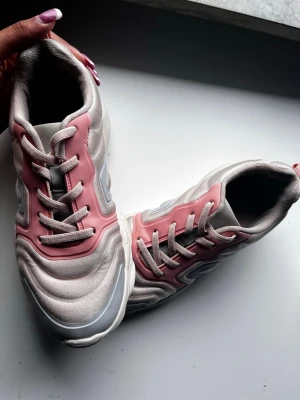 Sneakers i ljusrosa och grå - Säljer ett par snygga sneakers med vågig design i ljusrosa och grå toner. Skorna har platt sula, rund tå och snörning. Materialet är syntet med detaljer i rosa och grått, samt vit sula med mönstrad struktur. Perfekt för dig som gillar sportig och modern stil.