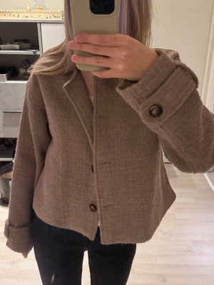 Beige kavajkappa med knappar - Säljer en beige kavajkappa med mjuk och varm textur och snygga bruna knappar framtill. Jackan har en rak, lite croppad passform och långa ärmar med dekorativa manschetter. Perfekt för dig som gillar stilrena och klassiska plagg med en modern twist.
