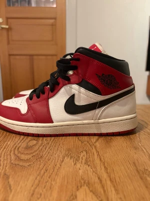 Nike Air Jordan 1 Mid Chicago US8 - Nike Air Jordan 1 Mid Chicago i klassisk röd, vit och svart färg. Skorna har snörning, rund tå och platt sula. Ikonisk Jumpman-logga på tungan och Air Jordan-märke på sidan. Tillverkade i skinn med perforerad tåbox för extra ventilation. Perfekt för dig som gillar streetwear och sneakers.