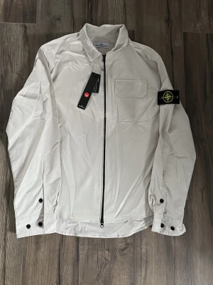 Vit vindjacka från Stone Island - Snygg vit vindjacka från Stone Island med klassisk kompasspatch på ärmen. Jackan har en rak passform, bröstficka, hel dragkedja framtill och knappar vid ärmslut. Materialet är lätt och känns fräscht, perfekt för vår och höst. En clean jacka med street-vibe.