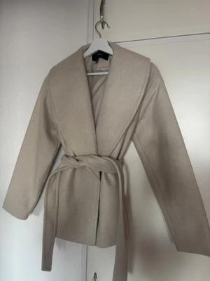Beige kappa med bälte från Vero Moda - Snygg beige kappa från Vero Moda med bred krage och knytbälte i midjan. Jackan har långärmade ärmar och en stilren, klassisk look som passar perfekt till höst och vinter. Materialet känns mjukt och har en ull-liknande finish. Köpte denna jacka för 900kr och aldrig använd.