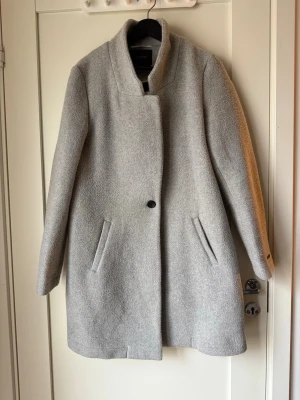 Ljusgrå kappa från Scotch & Soda - Säljer en stilren ljusgrå kappa från Scotch & Soda. Kappan har rak passform, hög krage och dolda knappar framtill. Tillverkad i en mjuk mix av ull och polyester som ger en skön och varm känsla. Två snedställda fickor framtill och enkel, clean look. Storlek 4 ca storlek XS/S enligt scotch soda storleks guide 