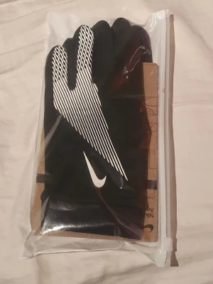 Nike Academy svarta fotbollshandskar - Säljer ett par svarta Nike Academy fotbollshandskar med vitt Swoosh-tryck och coola vita linjer på ovansidan. Handskarna har greppdetaljer i handflatan och är tillverkade i ett stretchigt syntetmaterial som håller händerna varma under träning eller match. STORLEK M-L