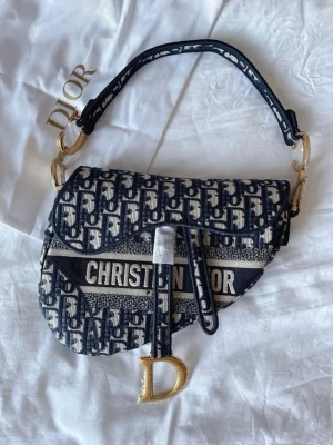 Dior Saddle Bag i blått mönster - Säljer en ikonisk Dior Saddle Bag med blått och vitt monogrammönster. Väskan har guldfärgade metalldetaljer, inklusive ett stort D-hänge, och ett brett axelband. Materialet är kraftig bomullscanvas med tydlig Dior-logga framtill. Perfekt statement piece för din outfit.