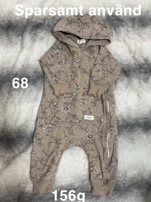 Blommig byxdress från Newbie - Supersöt brun byxdress med blommigt mönster från Newbie. Har huva, långa ärmar och muddar vid ärmslut och benslut. Dragkedja längs sidan gör den enkel att ta på och av. Perfekt för mysiga dagar.🌸 Sparsamt använd. Storlek 68