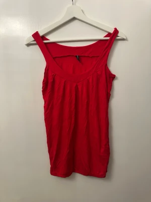 Röd ärmlös linnetopp - Snygg röd linnetopp med breda axelband och djup rundad ringning. Toppen är enkel och stilren, perfekt att matcha med jeans eller kjol. Tillverkad i mjuk bomull som känns skön mot huden.