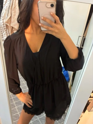 Svart byxdress med spetsdetalj - Säljer en svart byxdress med långa ärmar och omlott-ringning. Byxdressen har resår i midjan och shorts med fina spetsdetaljer längst ner. 