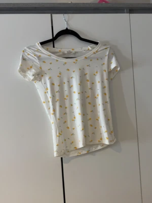 Vit blommig topp från 157 XS - Använd 1 gång.Supersöt vit topp från 157 i storlek XS, täckt med små gula blommor och gröna blad. Modellen har rund halsringning och korta ärmar. Perfekt till sommaren och enkel att matcha med jeans eller kjol.