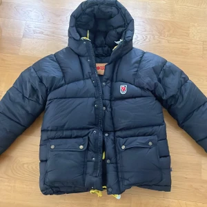 Svart dunjacka från Fjällräven - Säljer en svart, tjock dunjacka från Fjällräven med huva och gula snören. Jackan har två stora fickor med knappar framtill, dragkedja och Fjällräven-logga på bröstet. Perfekt för kalla dagar och har en riktigt schysst puffer-look. Jag är 1,55 så den är ganska stor på mig, skulle säga att den passar över 1,65. Den är ganska bred också. Passar som XS.