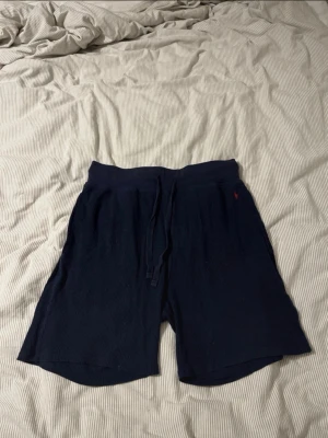 Mörkblå shorts från Polo Ralph Lauren - Säljer ett par mörkblå shorts från Polo Ralph Lauren med snörning i midjan och röd broderad logga på vänster sida. Shortsen är ribbade och gjorda i mjuk bomullsmix, perfekta för chill dagar. Klassisk loose passform och midwaist.