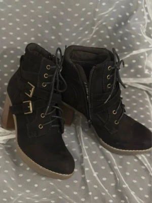 Svarta Attitude boots med klack och snörning 38,  - Säljer ett par svarta boots med grov klack, snörning och dragkedja på sidan. Skorna har två dekorativa spännen i guld och är gjorda i mocka-look. Rund tå och platt sula framtill. Perfekta för dig som gillar lite edgy stil.