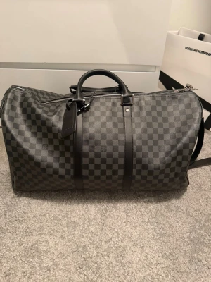 Louis Vuitton Damier Graphite resväska - Säljer en snygg Louis Vuitton resväska i Damier Graphite-mönster. Väskan är svart och grå med klassiskt rutigt mönster, har detaljer i skinn och silverfärgad dragkedja med hänglås. Perfekt storlek för weekendresor och har både handtag och axelrem.
